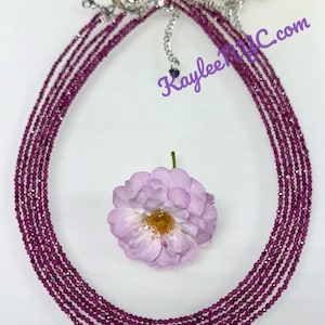 May include: A multi-strand necklace with small, faceted, deep pink beads. The necklace has a silver-colored clasp and is displayed with a light purple flower. The text "KayleeNYC.com" is visible in the image.