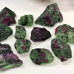 Pode incluir: Uma coleção de cristais brutos de zoisite com inclusões de rubi. As pedras exibem uma mistura de tons verdes, pretos e vermelhos profundos. Os cristais variam em tamanho e forma, mostrando texturas e padrões naturais.
