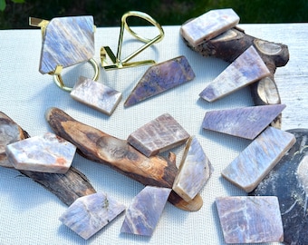 May include: An assortment of polished, rectangular and geometric-shaped stones in shades of lavender, white, and gray, arranged with a gold-colored ring and decorative metal stand. The stones are displayed on a white surface with natural wood accents.