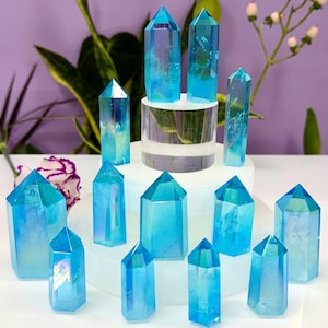 May include: An assortment of iridescent blue crystals in various shapes and sizes. Some crystals are tall and pointed, while others are shorter with flat tops. The crystals are arranged on a white surface with a purple background and greenery.