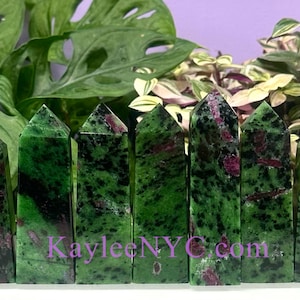 May include: A collection of 10 green and red ruby zoisite crystal points. The points are arranged in a row on a white surface. The points are all about the same size and shape. The points are a beautiful green color with red inclusions.