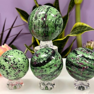 Könnte beinhalten: Vier polierte Zoisitkugeln, jeweils auf einem transparenten Acrylständer. Die Kugeln zeigen eine grüne Farbe mit schwarzen und roten Einschlüssen. Die Anordnung umfasst eine große Kugel und drei kleinere.