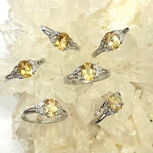 May include: Five silver rings with oval-shaped yellow gemstones set in a leaf design. The rings are displayed on a white crystal background.