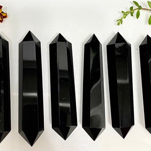 May include: Six polished black obsidian crystals, each shaped as a double-terminated wand with faceted sides and pointed ends. The stones are arranged in a vertical row, displaying a smooth, reflective surface and a deep, opaque black color.