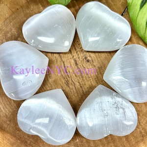 May include: Seven heart-shaped selenite stones arranged in a circular pattern. The stones are a translucent white color and have a smooth, polished surface. The background is a wooden surface with green leaves.