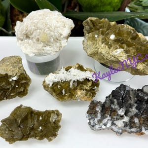 May include: A collection of various raw mineral specimens. The assortment includes a large, fluffy white mineral, several green and brown layered minerals, and a black and white mineral cluster. The minerals are displayed on a white surface.