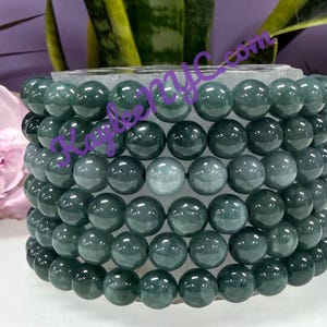 May include: A stack of jade bead bracelets in varying shades of green. The beads are round and smooth, with a polished finish. The bracelets are displayed on a clear, rectangular base. A pink flower and greenery are in the background.