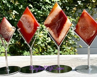 May include: Four diamond-shaped agate specimens in shades of red, orange, and white are displayed on silver metal stands. The agate pieces have unique internal patterns and textures, and are mounted on circular bases.