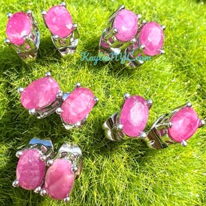 May include: Several silver rings with large, oval, pink gemstones are displayed. The gemstones are set in a four-prong setting. The rings are arranged on a bed of green grass. The image is well-lit, highlighting the gemstones' color and clarity.