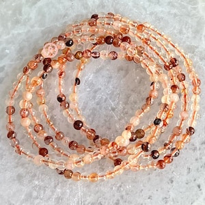 Op de afbeelding: Een set van zes delicate armbanden gemaakt met kleine, doorschijnende kralen in tinten roze, bruin en helder. De kralen hebben een subtiele glans en zijn geregen op een helder elastiek.