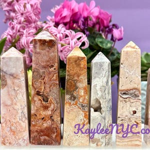 May include: Seven polished agate crystal points in various shades of pink, brown, and white. The points are arranged in a row on a white surface.