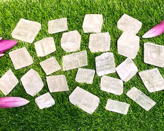 May include: A collection of white selenite crystals, some with a translucent appearance, arranged in a semi-circle on a green grassy surface. The crystals are various sizes and shapes, with some appearing more rectangular and others more cube-shaped.