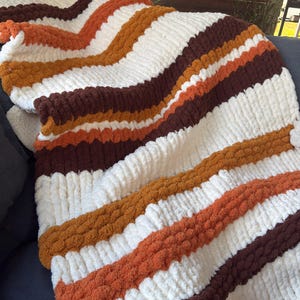 Puede incluir: Una manta de punto grueso con un patrón de rayas repetitivas en color crema, naranja quemado y marrón oscuro. La manta está confeccionada con bucles de hilo gruesos y suaves, creando una superficie afelpada y muy texturizada.