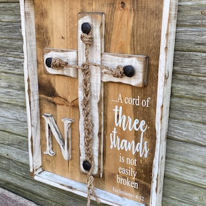 May include: Wooden cross wall art with a white-painted frame. The cross is made of wood with a rope design and the text "...A cord of three strands is not easily broken" from Ecclesiastes 4:12. The letter "N" is also visible.