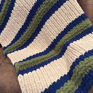 May include: A chunky knit blanket with alternating stripes of beige, navy blue, and olive green. The thick yarn creates a textured, cozy appearance. This handmade blanket is perfect for adding warmth and style to any living space.