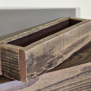 May include: A rustic wooden planter box with a natural wood finish. The planter has a rectangular shape and is made of reclaimed wood.