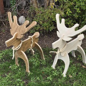 May include: Two wooden reindeer decorations, one natural wood color and one painted white, standing on a grassy lawn. Both reindeer have rope tied around their necks.