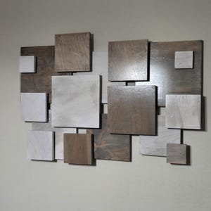 Puede incluir: Arte mural abstracto de madera compuesto por varios bloques cuadrados y rectangulares en tonos marrón, gris y blanco. Los bloques están dispuestos en un patrón asimétrico y superpuesto, creando un diseño geométrico moderno.