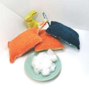 May include: Four colourful bath scrubbers made of terry cloth with a floral pattern. The scrubbers are in shades of orange, teal, and blue. A small white bowl filled with white cotton balls is in the foreground.