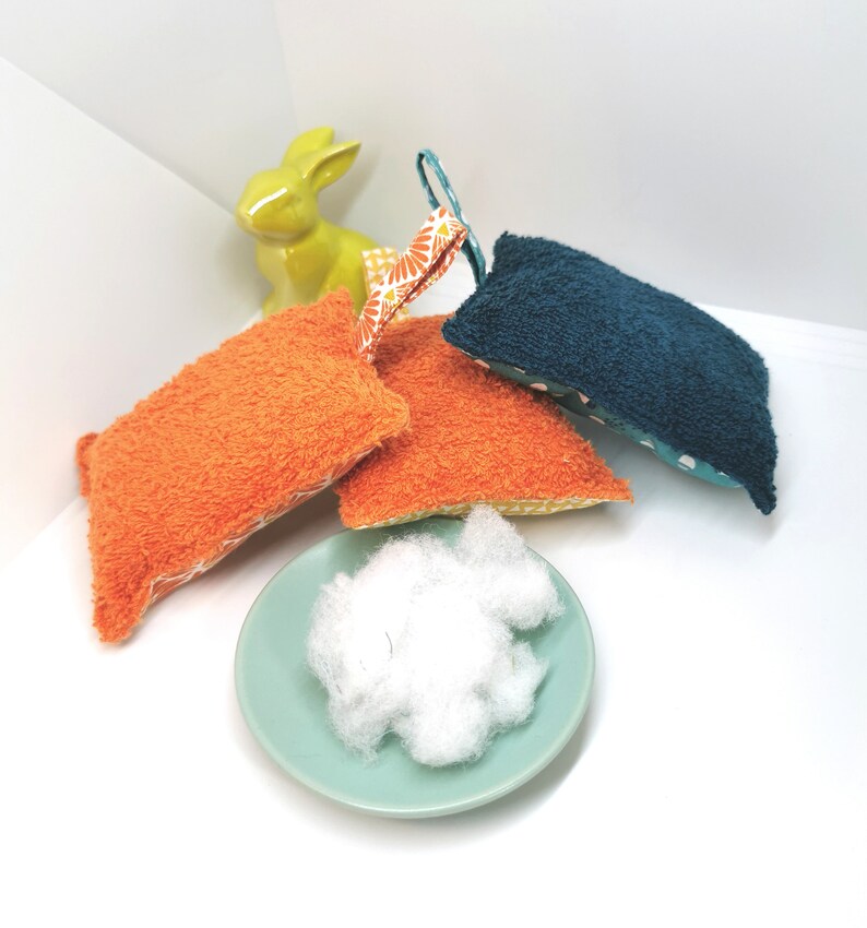 May include: Four colourful bath scrubbers made of terry cloth with a floral pattern. The scrubbers are in shades of orange, teal, and blue. A small white bowl filled with white cotton balls is in the foreground.