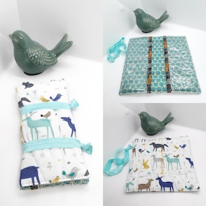 May include: A quilted fabric roll and a square fabric pouch featuring a white background with a pattern of blue, green, and yellow animals including deer, foxes, and birds, secured with light blue elastic bands and decorative beads.