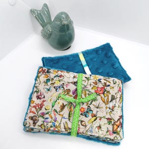 May include: A stack of rectangular fabric cloths featuring a cream background with a colourful botanical and butterfly print, backed with a textured teal dimpled fabric. The cloths are tied together with a bright green ribbon.