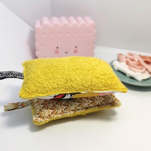 May include: A yellow and white fabric scrubby with a black and white patterned handle. The scrubby is made of two layers of fabric, one yellow and one white with a floral pattern.