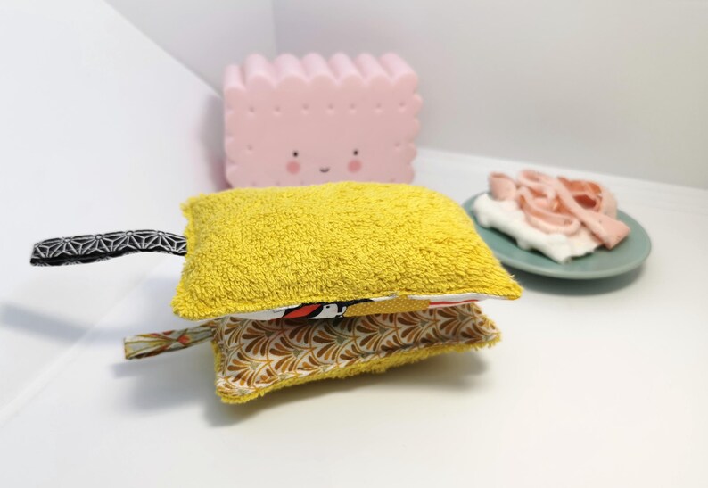 May include: A yellow and white fabric scrubby with a black and white patterned handle. The scrubby is made of two layers of fabric, one yellow and one white with a floral pattern.