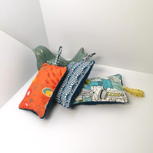 May include: Three colourful dishcloths with a teal border. The dishcloths have different patterns: orange with floral designs, white with blue geometric patterns, and white with a blue, green and yellow animal print.