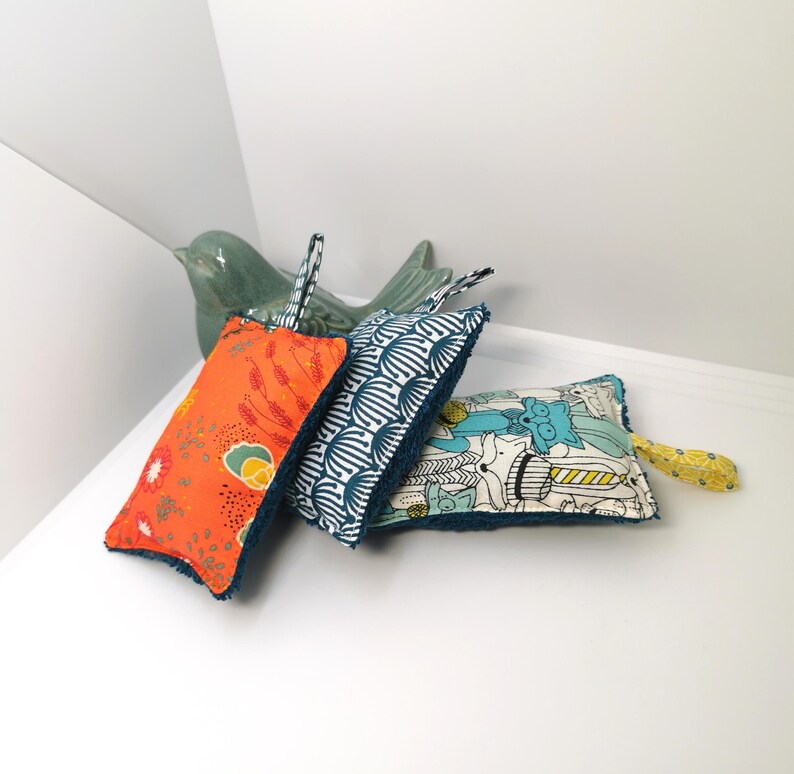 May include: Three colourful dishcloths with a teal border. The dishcloths have different patterns: orange with floral designs, white with blue geometric patterns, and white with a blue, green and yellow animal print.