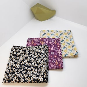 May include: Three fabric pocket squares with different floral patterns. The first square is black with white flowers, the second is purple with pink flowers and the third is beige with yellow flowers.