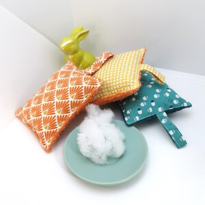 May include: Four colourful fabric squares with different patterns, including orange and white, yellow and white, and blue and white. The squares are arranged on a white surface with a small white fluffy object in the centre.