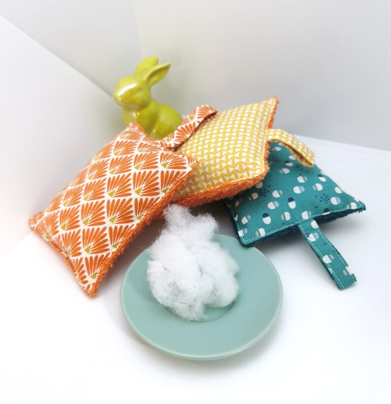 May include: Four colourful fabric squares with different patterns, including orange and white, yellow and white, and blue and white. The squares are arranged on a white surface with a small white fluffy object in the centre.