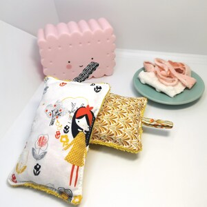 May include: Two colourful fabric scrubbers with yellow trim. One scrubber features a girl with red hair and a yellow dress in a white and grey floral pattern. The other scrubber has a yellow and gold geometric pattern.