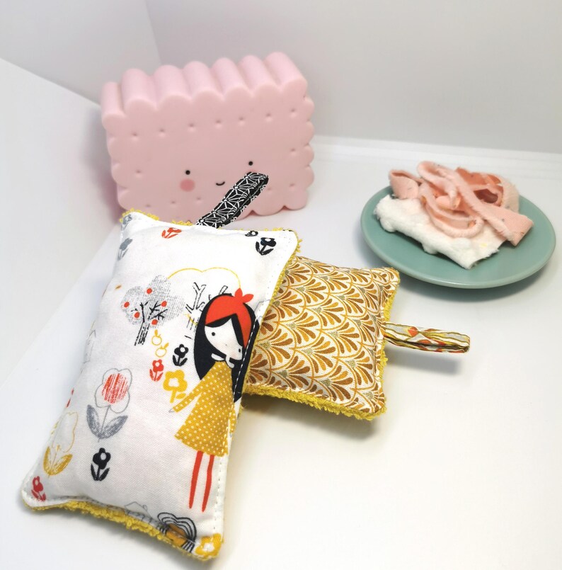 May include: Two colourful fabric scrubbers with yellow trim. One scrubber features a girl with red hair and a yellow dress in a white and grey floral pattern. The other scrubber has a yellow and gold geometric pattern.