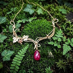 May include: A gold-toned necklace with a vine and leaf design, featuring a teardrop-shaped, deep red gemstone pendant. The necklace is set against a backdrop of green foliage and moss, creating a natural aesthetic.