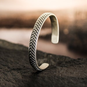May include: A silver cuff bracelet with a textured, scale-like pattern. The open-ended bracelet is displayed on a dark, textured surface, with a blurred background of a body of water and a sunset.