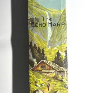 Könnte beinhalten: Ein rechteckiger Pappkarton für das Musikinstrument The Echo Harp. Die Schachtel zeigt eine farbenfrohe Illustration einer Berglandschaft mit einem Wasserfall, einer Holzhütte und einer Person auf einem Pfad. Text: The Echo Harp und M.Hohner.