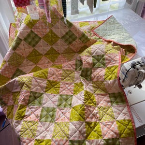 Pu&ograve; includere: Una trapunta fatta a mano con un design patchwork nei toni del rosa e del verde. La trapunta presenta un motivo a rombi con varie stampe floreali e geometriche. La trapunta &egrave; rifinita con un bordo rosa e ha un'etichetta che dice "MEESH QUILTS".