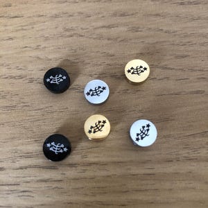 May include: Six round metal buttons featuring a stylised plant design with five stars surrounding it. The buttons are available in three colours: gold, silver, and black.