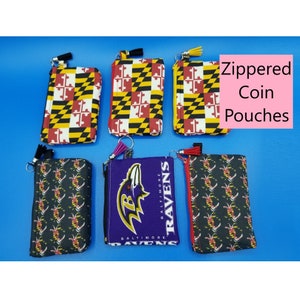 Może przedstawiać: Sześć zapinanych na zamek błyskawiczny portfeli na monety z różnymi wzorami. Dwa portfele przedstawiają flagę stanu Maryland, dwa portfele przedstawiają wzór czarno-biały, a dwa portfele przedstawiają logo Baltimore Ravens.