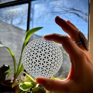 May include: A white glass sphere with a geometric pattern of overlapping circles and triangles. The sphere is being held in a hand.