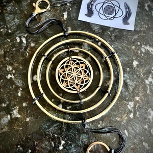 May include: A gold metal mobile with three concentric circles and a flower of life design in the center. The mobile is suspended from a black cord with a silver clip.