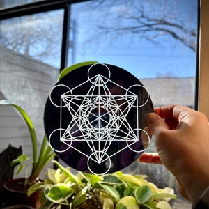 May include: A black circular mirror with a white geometric design, resembling a star with overlapping circles. The mirror is being held by a hand in front of a window.