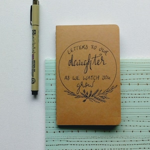 May include: A brown notebook with the text "Letters to our daughter as we watch you grow" written in black cursive. The notebook is sitting on a blue and white patterned surface.