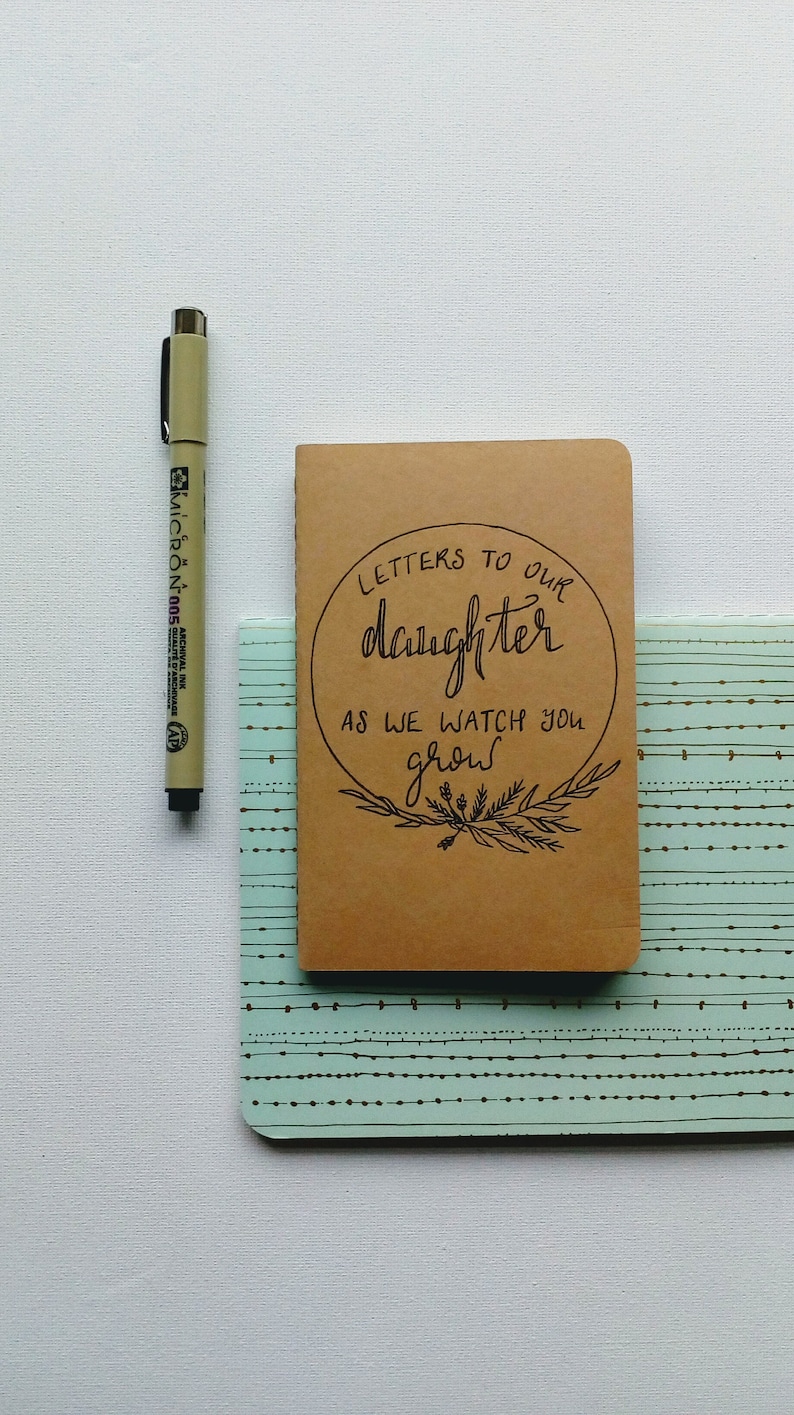 May include: A brown notebook with the text "Letters to our daughter as we watch you grow" written in black cursive. The notebook is sitting on a blue and white patterned surface.