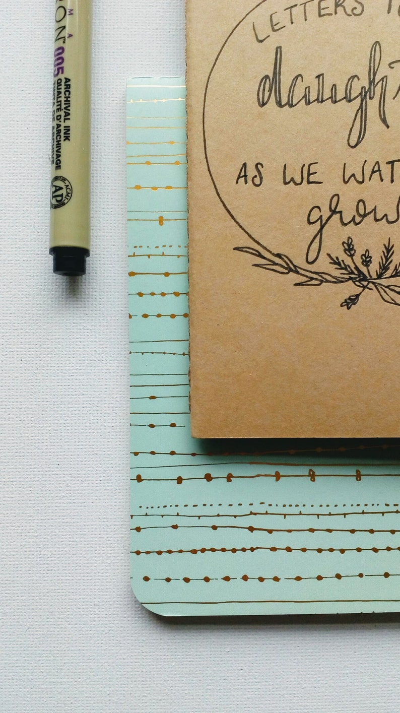 May include: A brown notebook with a light blue patterned cover. The notebook has a circle on the front with the text "Letters to my daughter as we grow".