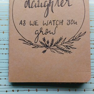 May include: A brown notebook with a circle on the front that says "Letters to our daughter as we watch you grow". The circle is surrounded by a wreath of leaves and flowers.