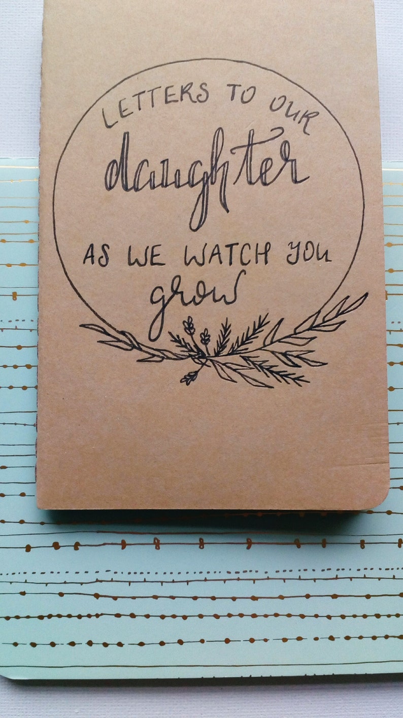 May include: A brown notebook with a circle on the front that says "Letters to our daughter as we watch you grow". The circle is surrounded by a wreath of leaves and flowers.