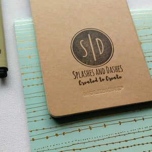 May include: A brown Moleskine notebook with a black circle logo that says "S|D" and the text "Splashes and Dashes Created to Create". The notebook is on top of a teal notebook with a gold pattern.